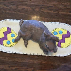 Puede incluir: Una figurita de conejo marrón tumbada sobre un camino de mesa de fieltro blanco con huevos de Pascua amarillos y morados.
