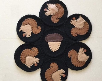May include: A felt wall hanging with a brown acorn in the center surrounded by six black circles with brown squirrels holding acorns.
