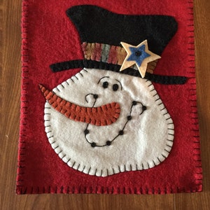 May include: A felt snowman ornament with a black top hat, a red nose, and black eyes. The snowman is on a red felt background with black stitching around the edges.