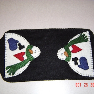 May include: A black felt table runner with two white felt snowmen appliques. Each snowman has a green scarf, red heart, and blue hat. The text "OCT 25 2011" is in the bottom right corner.