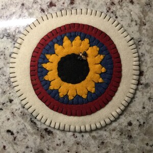 Pode incluir: Um descanso de panela redondo de feltro com um design de girassol em amarelo, vermelho, azul e preto. Duas pequenas abelhas estão no centro do girassol.