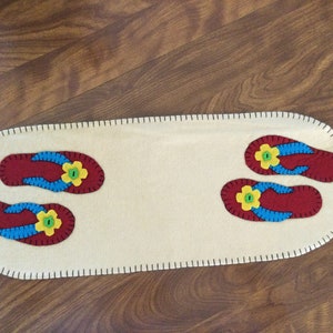 May include: A white felt table runner with a stitched border features two pairs of red, blue, and yellow felt flip-flops.