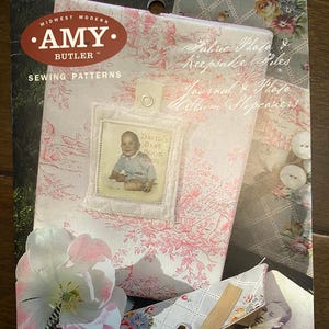 May include: A collection of fabric-covered memory keepers, including a journal and photo album slipcovers. The items feature floral and toile patterns in pink and white. One album displays a baby photo with the text "DAVID'S BABY BOOK 2002". The Amy Butler Sewing Patterns logo is visible.