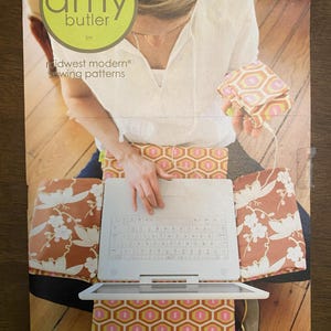 May include: A laptop cover and MP3 case set featuring a geometric pattern in shades of pink, orange, and white. The set includes a white laptop and a matching case. The Amy Butler logo is visible.