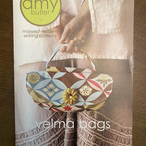 May include: A sewing pattern for "Velma Bags" by Amy Butler. The bag has a geometric floral pattern in brown, blue, yellow, and red. The pattern includes instructions for a handbag and shoulder bag.