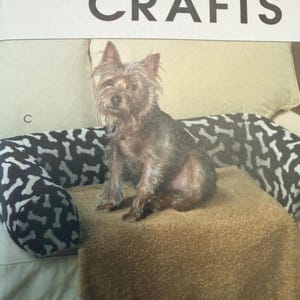 May include: A McCall's Crafts pattern cover featuring a small terrier dog sitting on a tan, textured pet bed. The bed has a black bolster with a white bone pattern. The letter C is printed on the left side of the image, and a small dog is shown at the bottom.