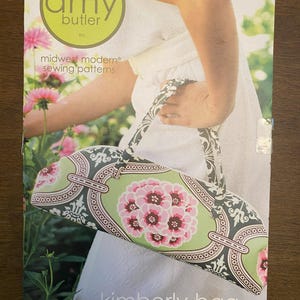 May include: A sewing pattern package featuring an Amy Butler design. The package displays a Kimberly bag with a floral pattern in pink and green. The bag has a black and white patterned handle. The text "midwest modern sewing patterns" is visible.