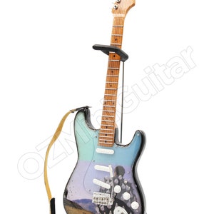 May include: A miniature electric guitar with a light blue, purple, and black body, resting on a black stand. The guitar features a landscape design and the text "Delicate Sound of Thunder". A tan strap is attached.