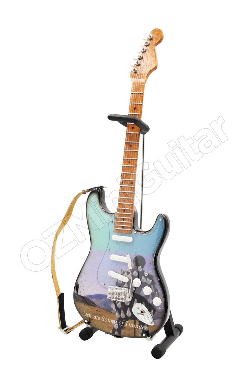 May include: A miniature electric guitar with a light blue, purple, and black body, resting on a black stand. The guitar features a landscape design and the text "Delicate Sound of Thunder". A tan strap is attached.