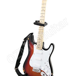 Może przedstawiać: Elektryczna gitara w kolorze sunburst z białym pickguardem, czarnym osprzętem i czarnym paskiem. Gitara stoi na czarnym stojaku.