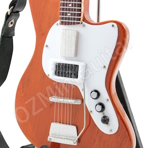 May include: A miniature replica of a vintage electric guitar with a white pickguard, a brown body, and a black strap. The guitar is on a black stand.