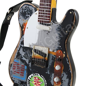 Może przedstawiać: Czarna gitara elektryczna z białym pickguadem i zużytym, postarzanym wykończeniem. Gitara ma skórzany pasek i jest ozdobiona naklejkami, w tym jedną z napisem "Trash City".