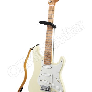 May include: A white electric guitar with a brown wooden neck and a black strap. The guitar is on a black stand.