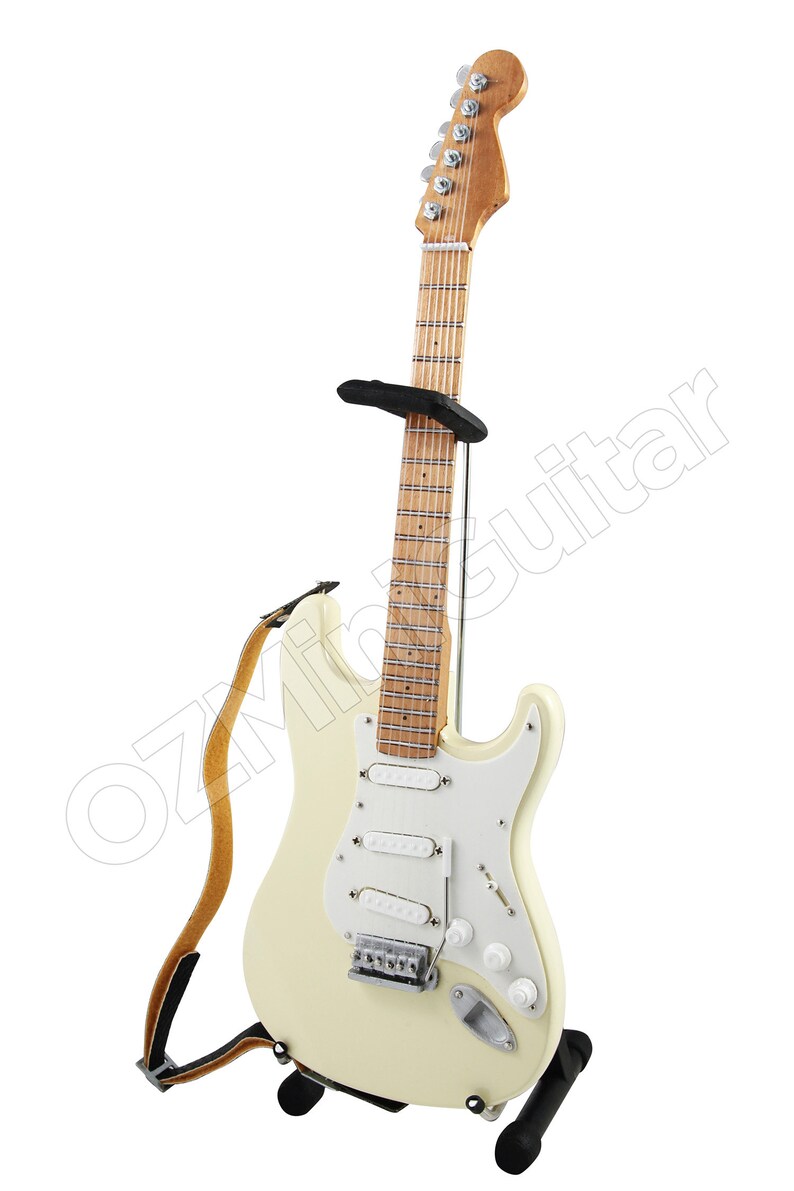 May include: A white electric guitar with a brown wooden neck and a black strap. The guitar is on a black stand.