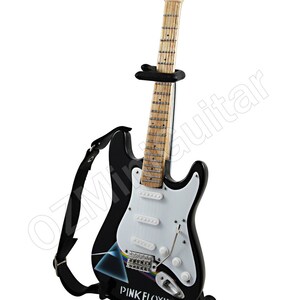 Pu&ograve; includere: Una chitarra elettrica nera con un battipenna bianco e un logo Pink Floyd sul corpo. La chitarra &egrave; su un supporto nero e ha una tracolla nera.