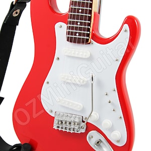 Puede incluir: Una guitarra el&eacute;ctrica roja con una golpeador blanco y herrajes cromados. La guitarra tiene una correa y est&aacute; en un soporte.