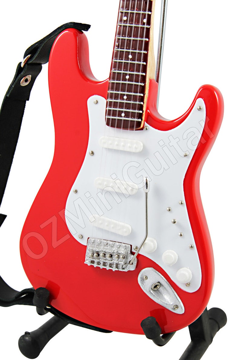 Puede incluir: Una guitarra el&eacute;ctrica roja con una golpeador blanco y herrajes cromados. La guitarra tiene una correa y est&aacute; en un soporte.