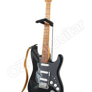 May include: A black electric guitar with a white pickguard and a woman's image on the body. The guitar is on a stand with a brown strap.