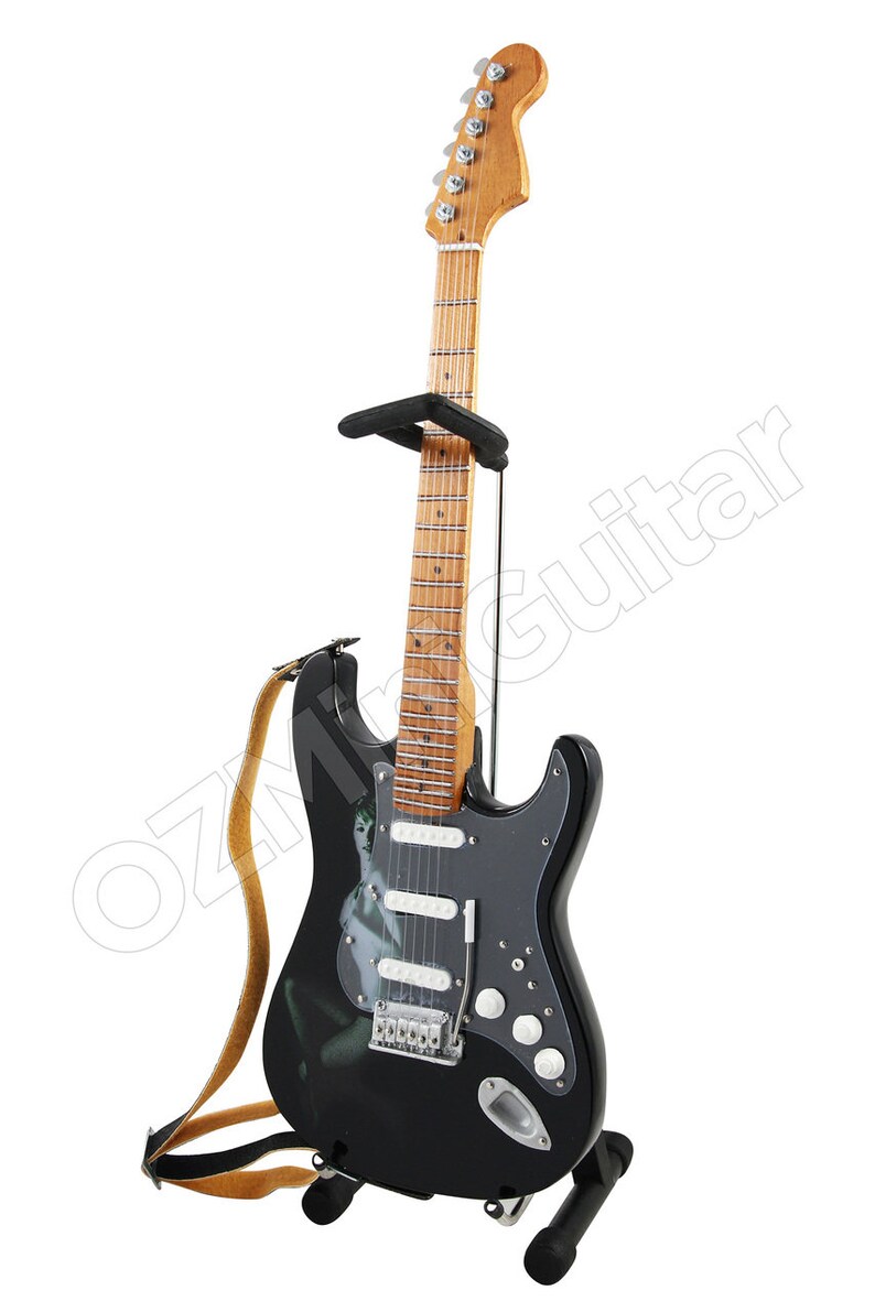 May include: A black electric guitar with a white pickguard and a woman's image on the body. The guitar is on a stand with a brown strap.