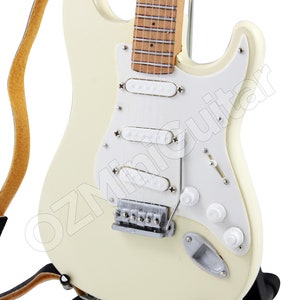 May include: A white electric guitar with a brown wooden fretboard and a black strap. The guitar is on a black stand.