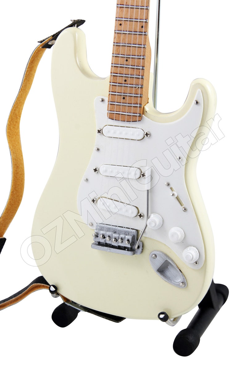 May include: A white electric guitar with a brown wooden fretboard and a black strap. The guitar is on a black stand.