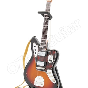 May include: A miniature electric guitar with a sunburst finish, featuring a tortoiseshell pickguard, silver hardware, and a light tan strap. The instrument is displayed on a black metal stand, with the text OZGuitar visible across the image.
