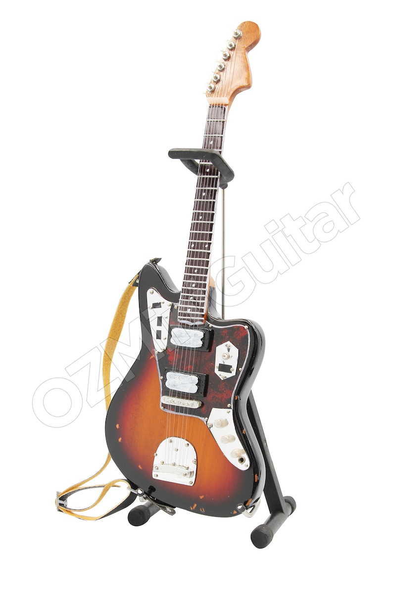 May include: A miniature electric guitar with a sunburst finish, featuring a tortoiseshell pickguard, silver hardware, and a light tan strap. The instrument is displayed on a black metal stand, with the text OZGuitar visible across the image.