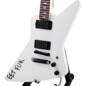 Puede incluir: Una guitarra eléctrica blanca con una distintiva forma de cuerpo Explorer, apoyada en un soporte negro. La guitarra presenta pastillas negras, perillas y el texto "EET FUK" en el cuerpo.