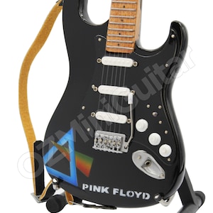 May include: A black electric guitar with a white pickguard and a blue and rainbow triangle graphic. The guitar has a brown wooden neck and is on a black stand. The text "PINK FLOYD" is printed on the body of the guitar.