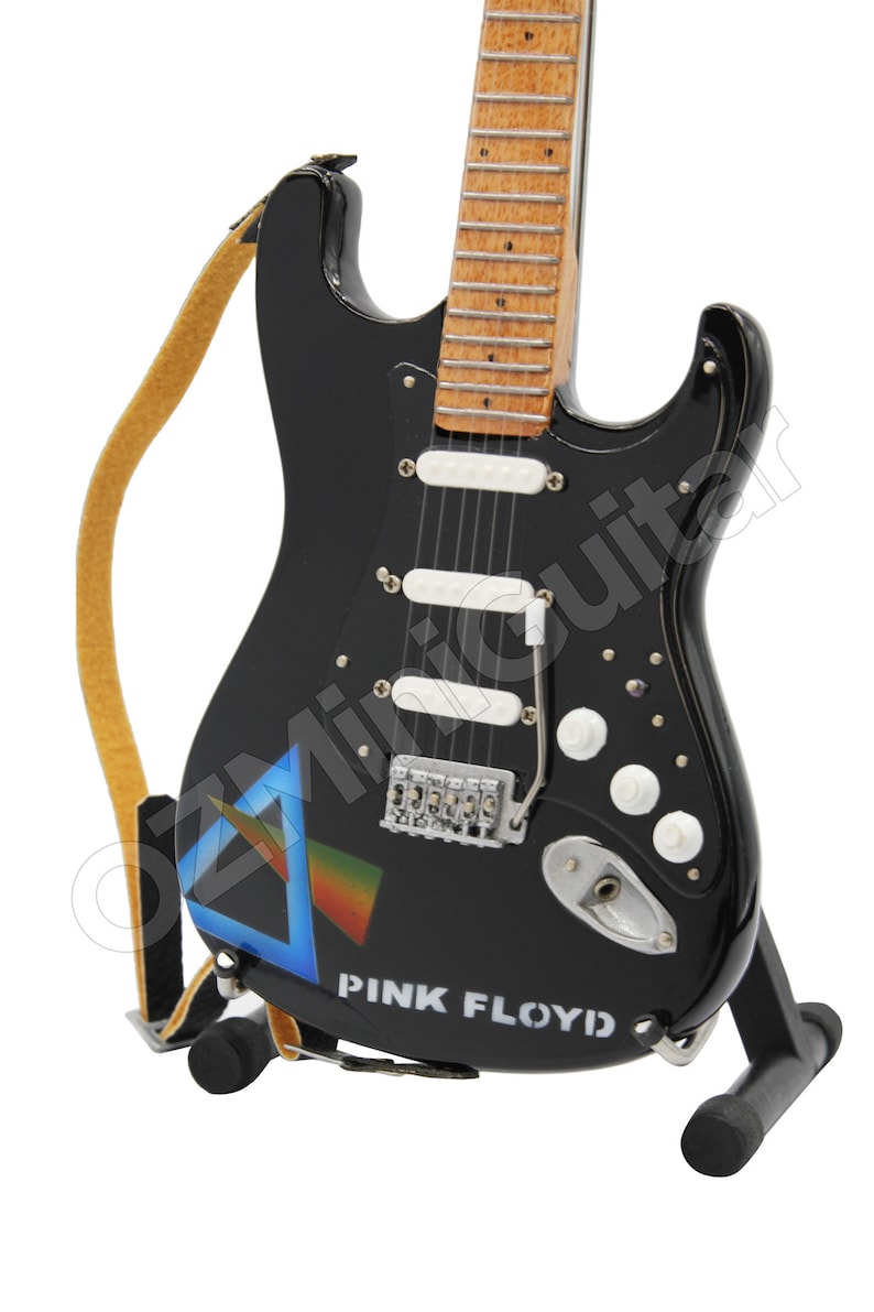 May include: A black electric guitar with a white pickguard and a blue and rainbow triangle graphic. The guitar has a brown wooden neck and is on a black stand. The text "PINK FLOYD" is printed on the body of the guitar.
