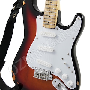 Może przedstawiać: Miniaturowa gitara elektryczna z wykończeniem sunburst, białym pickguardem i czarnym paskiem. Gitara stoi na czarnym stojaku.
