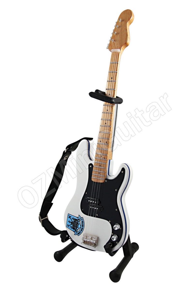 May include: White electric guitar with a black pickguard, blue trim, and a wooden neck. The guitar has a black strap and is displayed on a black stand. A logo is visible on the body.