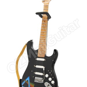 May include: Black electric guitar with a brown wooden neck and a white pickguard. The guitar has a strap and a stand. The guitar has a Pink Floyd logo and a graphic of a prism on the body.