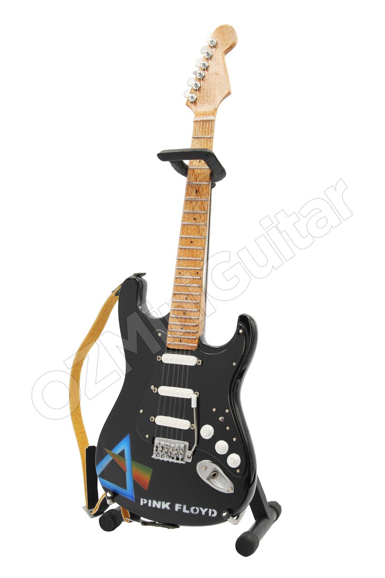May include: Black electric guitar with a brown wooden neck and a white pickguard. The guitar has a strap and a stand. The guitar has a Pink Floyd logo and a graphic of a prism on the body.