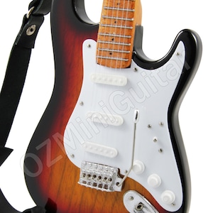 May include: A miniature electric guitar with a sunburst finish, black hardware, and a white pickguard. The guitar is on a black stand and has a black strap attached.