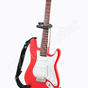 Puede incluir: Una guitarra el&eacute;ctrica roja con una golpeador blanco y una correa negra. La guitarra est&aacute; en un soporte negro.