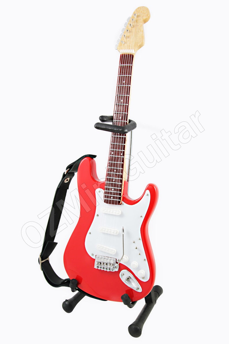 Puede incluir: Una guitarra el&eacute;ctrica roja con una golpeador blanco y una correa negra. La guitarra est&aacute; en un soporte negro.