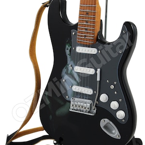May include: Black electric guitar with a white pickguard and a woman's image on the body. The guitar has a brown strap and is on a black stand.