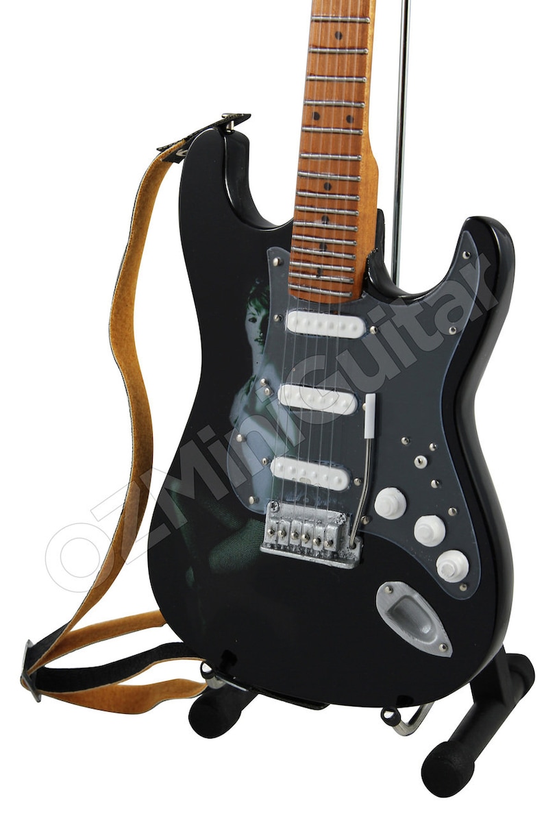 May include: Black electric guitar with a white pickguard and a woman's image on the body. The guitar has a brown strap and is on a black stand.