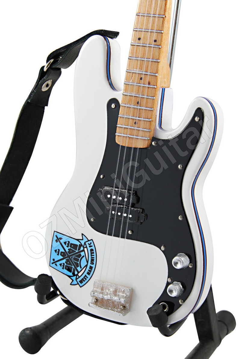 May include: A white electric guitar with a black pickguard and a West Ham United FC logo. The guitar has a black strap and is mounted on a black stand. The body is outlined with blue trim.
