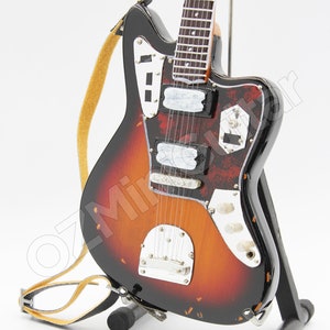 May include: A miniature electric guitar with a sunburst finish, featuring a black pickguard, chrome hardware, and a yellow strap. The instrument is mounted on a black stand and displays a detailed fretboard with white dot inlays and six strings.