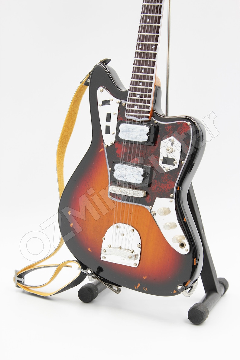 May include: A miniature electric guitar with a sunburst finish, featuring a black pickguard, chrome hardware, and a yellow strap. The instrument is mounted on a black stand and displays a detailed fretboard with white dot inlays and six strings.