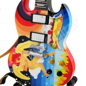May include: A miniature electric guitar with a colorful, psychedelic design. The body of the guitar is painted with a rainbow of colors, including red, orange, yellow, green, blue, and purple. The guitar also features a nude female figure with wings, a cloud, and stars. The guitar is on a black stand.
