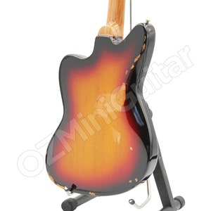 May include: A miniature electric guitar with a sunburst finish featuring a gradient from black to red to yellow. The body shows distressed edges with visible wear. It is mounted on a black metal display stand. The text OZMiniGuitar is visible.