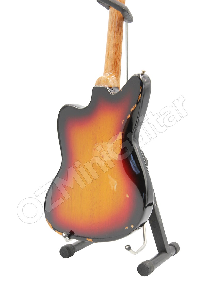 May include: A miniature electric guitar with a sunburst finish featuring a gradient from black to red to yellow. The body shows distressed edges with visible wear. It is mounted on a black metal display stand. The text OZMiniGuitar is visible.