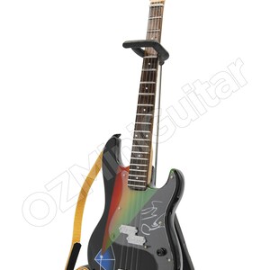 Puede incluir: Un bajo el&eacute;ctrico negro con pintura de colores arco&iacute;ris y una firma en el cuerpo. La guitarra est&aacute; en un soporte con una correa adjunta.