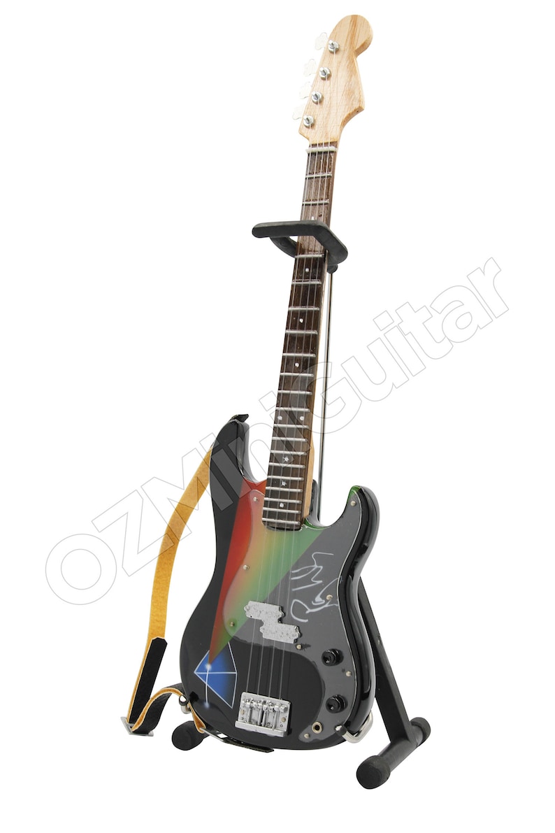 Puede incluir: Un bajo el&eacute;ctrico negro con pintura de colores arco&iacute;ris y una firma en el cuerpo. La guitarra est&aacute; en un soporte con una correa adjunta.