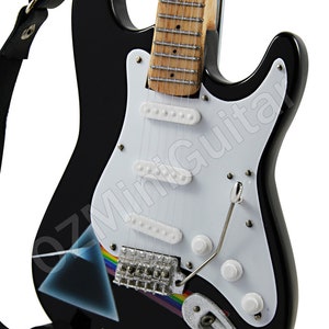 Pu&ograve; includere: Una chitarra elettrica nera con un battipenna bianco e un logo Pink Floyd sul corpo. La chitarra ha una tastiera in legno scuro e un ponte argentato. La chitarra &egrave; su un supporto nero.