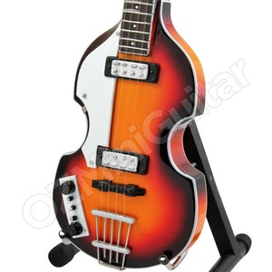 Op de afbeelding: Een rode, oranje en zwarte elektrische vioolbasgitaar met een witte pickguard. De gitaar staat op een zwarte standaard.