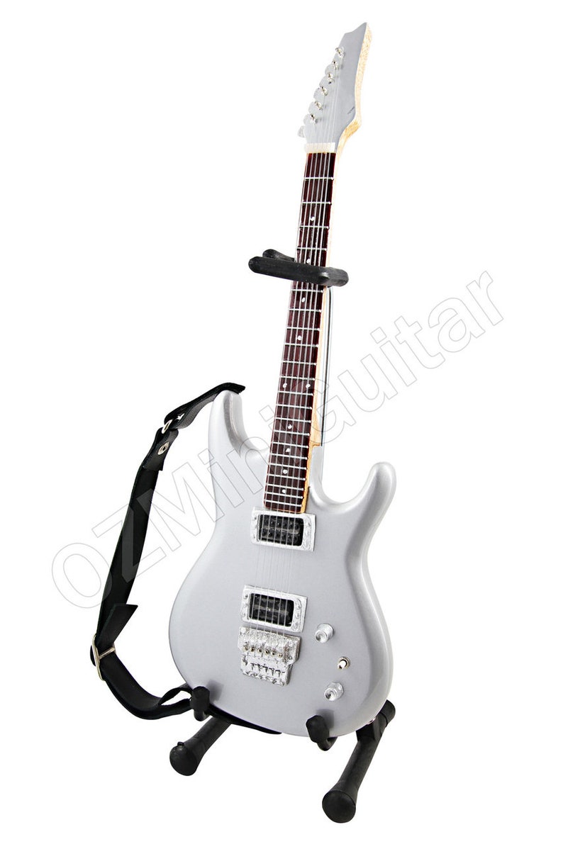 Puede incluir: Una guitarra el&eacute;ctrica plateada con una correa negra y un soporte negro. La guitarra tiene un diapas&oacute;n de palisandro y herrajes cromados.